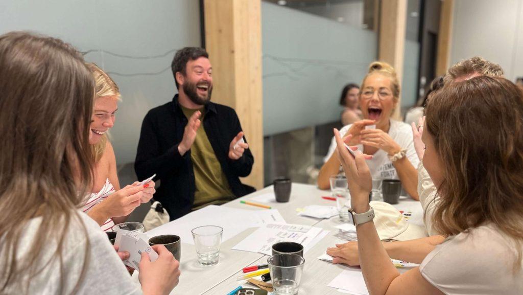 En grupp personer skrattar runt ett bord i en workshop.